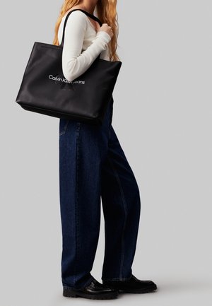 Černá taška přes rameno s logem ve stříbrném písmu, vyrobená z hladkého materiálu, s pevnými ušima a prostorným designem. Nositá s volnými džíny.