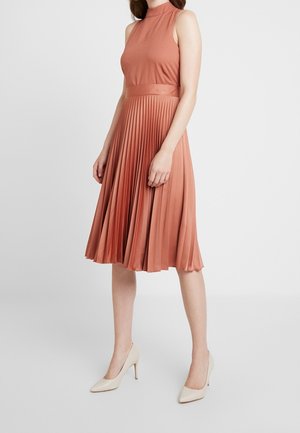 Vrouw draagt een mouwloze, roestkleurige geplooide midijurk met hoge hals en beige hoge hakken, staand op een witte achtergrond.