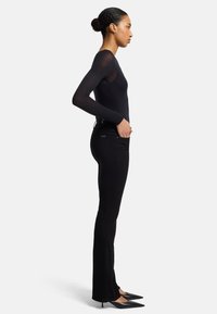 Svart langermet topp laget av lett stoff, kombinert med tettsittende svarte snekkerjeans. Modellen har på seg spisse svarte hæler, vist i profil.