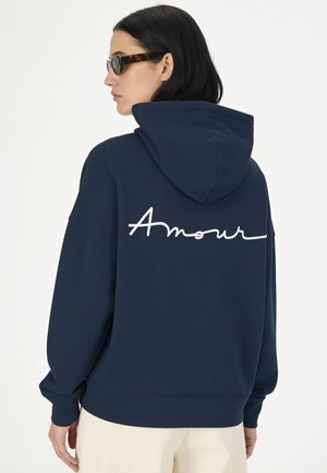 Frau mit dunklen Haaren, die Schildpatt-Sonnenbrillen und einen dunkelblauen Hoodie mit weißem "Amour"-Text auf dem Rücken trägt, von hinten.