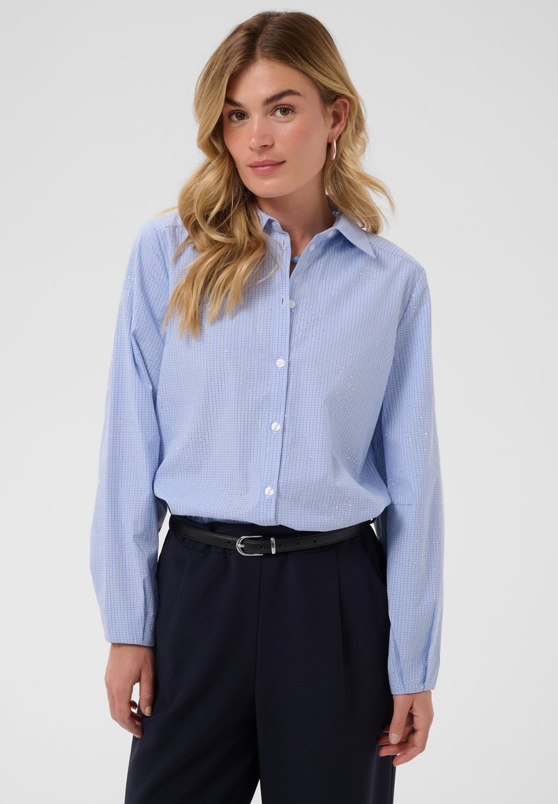 Femme aux longs cheveux blonds porte une chemise bleu clair à boutons et un pantalon foncé taille haute avec une ceinture, debout sur un fond uni.