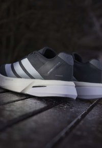 Par grå og hvide Adidas Adizero Boston 13 løbesko, der hviler på en træoverflade med sløret mørk baggrund.