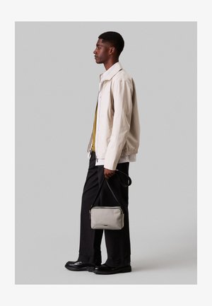 Casaco bege claro com bainha elástica, combinado com uma camisa verde e calças pretas. A portar uma pequena mala cinza a tiracolo. Sapatos pretos.