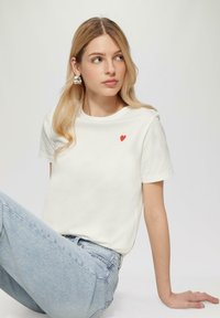 Witte katoenen t-shirt met korte mouwen, voorzien van een klein rood hartje op de linkerborst. Gecombineerd met lichtblauwe spijkerbroek.
