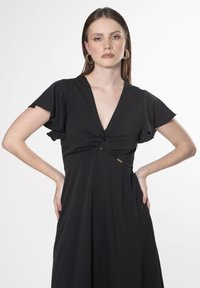 Vestido negro con escote en V profundo, mangas cortas y vaporosas, y detalle de nudo en el frente. Presenta una textura suave y un sutil acento de logo.