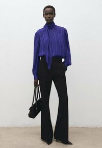 Blusa a righe viola con collo alto, pantaloni neri svasati e una borsa nera. L'outfit presenta texture lisce e una vestibilità su misura.