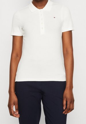 Personne portant un polo blanc ajusté à manches courtes avec boutons et petit logo, associé à un pantalon bleu marine foncé.