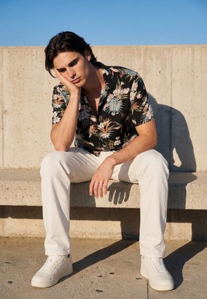 Młody mężczyzna w kwiecistej koszuli i białych spodniach siedzi na betonowej ławce, opierając głowę na dłoni, patrzy zamyślony na czyste niebieskie niebo.