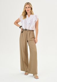 Blusa bianca a volant con bottoni, abbinata a pantaloni beige a gamba larga e una cintura intrecciata. Sandali con tacco alto e fasce completano l'outfit.
