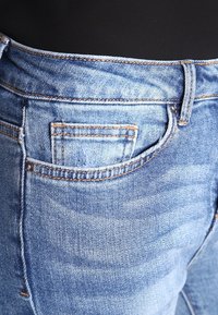 Vaate denim-farkut vaaleansinisessä värissä, joissa on kulutettu pinta, pieni takatasku ja oranssit ompeleet saumoissa.