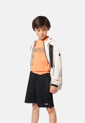 Veste blanche zippée en tissu texturé avec des accents noirs, t-shirt orange vif avec le logo "BOSS", shorts noirs avec des zips latéraux.