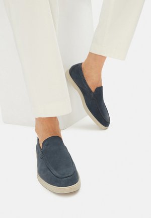 Námořnické semišové slip-on boty s kulatou špičkou, béžovou gumovou podrážkou, minimálním šitím a bohatým detailem loga, postavené na světlých kalhotách.