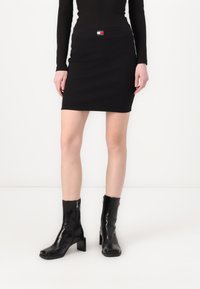 Svart bodycon skjørt med en liten logo-lapp i midjen, sammen med blanke svarte ankelstøvler med firkantet tå og blokkhel.