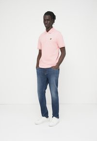 Άνδρας με πλεκτές κοτσίδες, ντυμένος με ροζ πόλο μπλούζα, γαλάζια τζιν και λευκά αθλητικά παπούτσια, στέκεται με τα χέρια στις τσέπες.