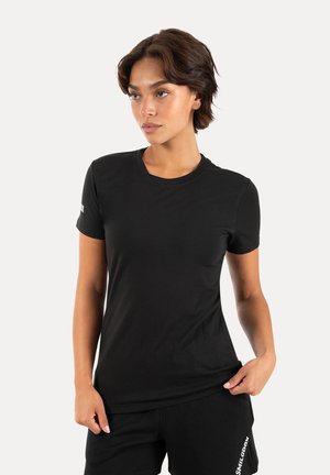 Frau mit kurzen braunen Haaren, die ein schlichtes schwarzes kurzärmeliges Shirt und schwarze Hose mit weißem Text am linken Bein vor weißem Hintergrund trägt.