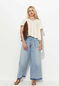 T-shirt crème à col en V et coupe courte associé à un jean large taille haute bleu clair et des sandales marron à clous, tenant un sac à franges marron.