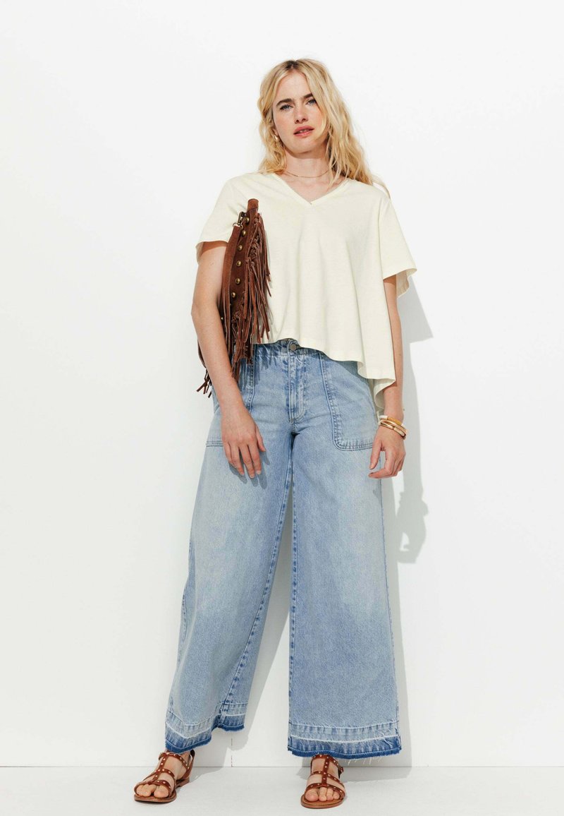 T-shirt crème à col en V et coupe courte associé à un jean large taille haute bleu clair et des sandales marron à clous, tenant un sac à franges marron.