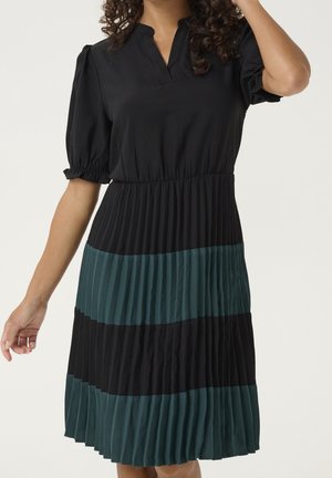 Vrouw draagt een zwarte jurk met korte pofmouwen en een geplooide rok met afwisselend brede zwarte en donkergroene horizontale strepen.