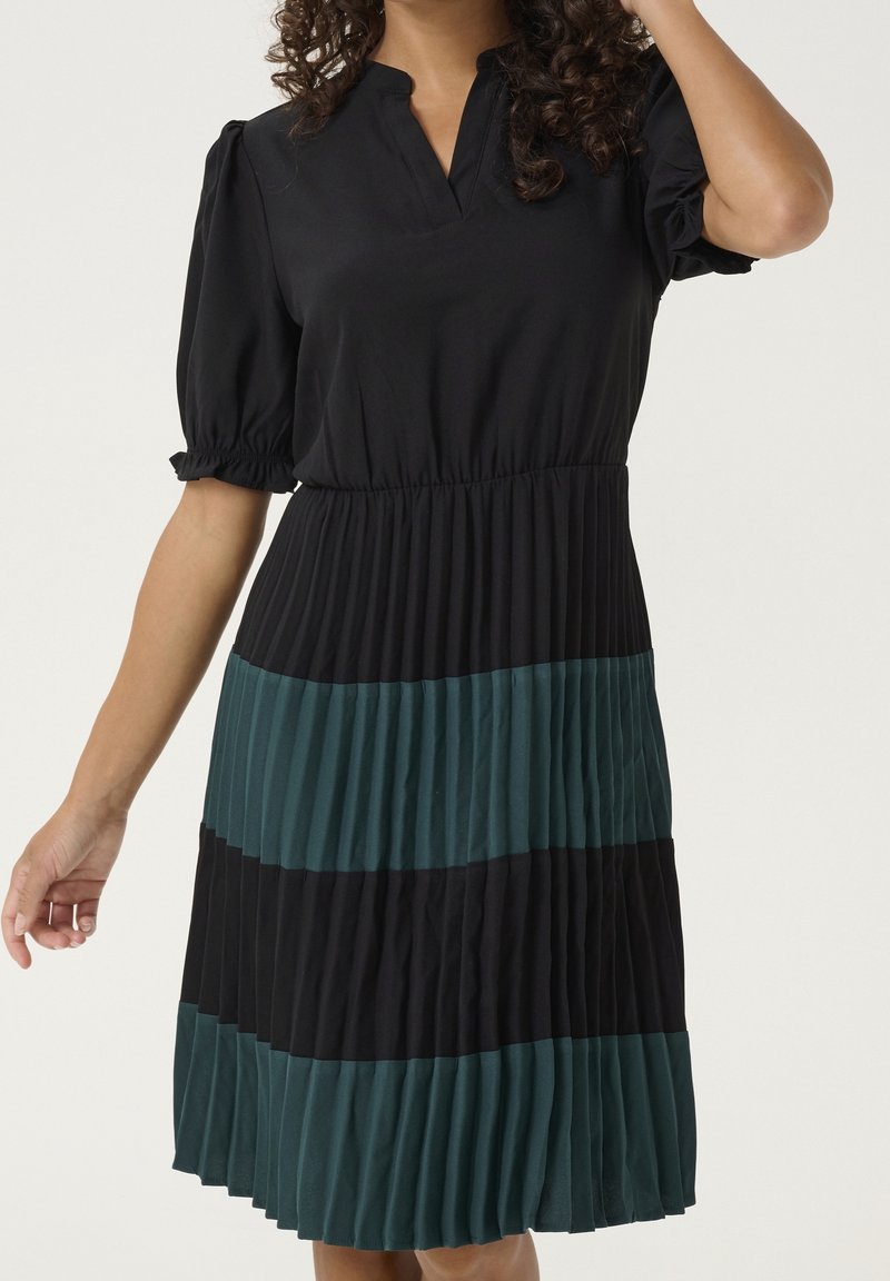 Femme portant une robe noire avec des manches courtes bouffantes et une jupe plissée comportant des larges rayures horizontales alternées noir et vert foncé.