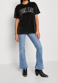 Persona vestida con una camiseta negra de "Tommy Jeans", jeans acampanados de color azul claro y botas de tacón negras que se apoya contra un fondo liso.