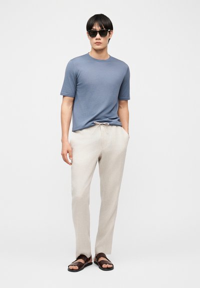 Άνδρας που φοράει μαύρα γυαλιά ηλίου, μπλε κοντομάνικο πουκάμισο, ανοιχτό μπεζ παντελόνι με κορδόνι και σκούρα καφέ σανδάλια, στέκεται με το ένα χέρι στην τσέπη.