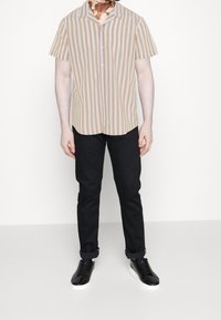 Muž oblečený v krátkém rukávu košile s béžovými a bílými vertikálními pruhy, černých kalhotách s vyhrnutými nohavicemi a černých teniskách s bílou podrážkou stojící na jednobarevném pozadí.