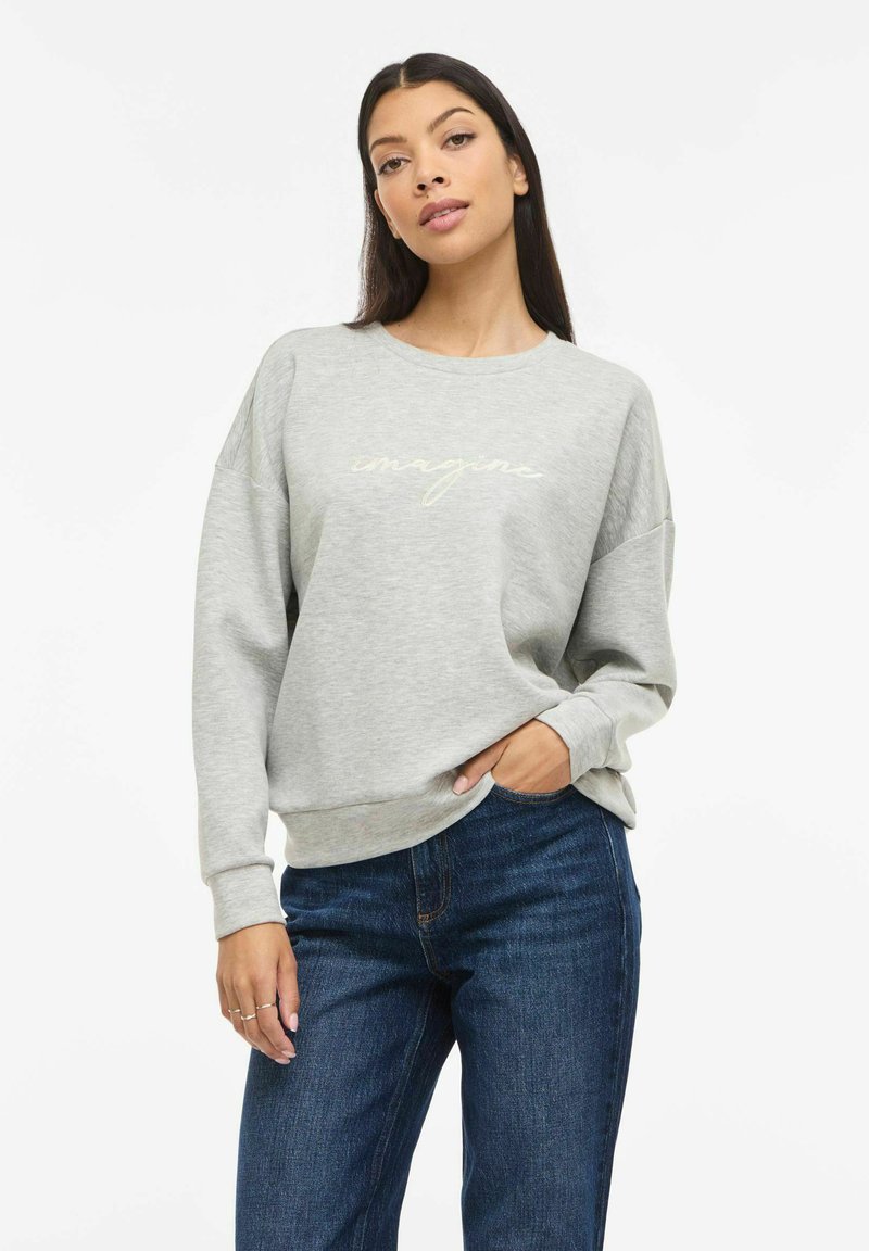 Lysegrå genser med rund hals, senkede skuldre og teksten "imagine" i en myk gul kursiv skrift. Kombinert med mørkeblå jeans.