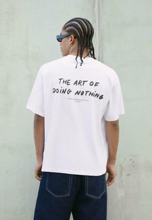 T-shirt bianco oversize con bold testo nero sul retro che recita "L'ARTE DI NON FARE NULLA", con scollo a girocollo e maniche corte.