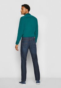 Chemise teal à manches longues, jean bleu foncé et chaussures navy. La chemise a un col ; le jean présente des poches arrière et des détails de couture.