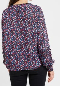 Femme portant un chemisier à manches longues avec un motif floral dense en rouge, bleu et blanc, associé à un pantalon noir, vue de dos.