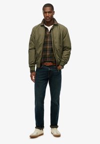 Olive green bomber jacket over a checkered flannel shirt, paired with dark jeans and cream sneakers. Brown belt and watch visible.