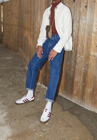 Vit snörjacka över en brun polotröja, blå uppvikta jeans och vita sneakers med röda ränder. Bakgrunden har en vägg av trä.