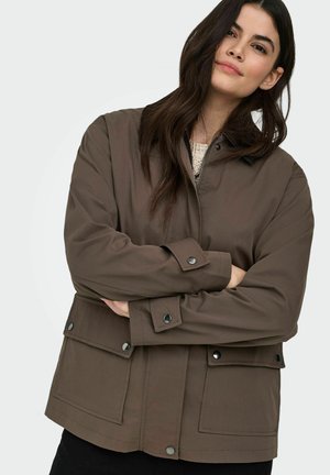 Hosszú sötét hajú nő barna, gombos, elülső zsebes dzsekit visel, karját keresztbe téve, egyszerű világos háttér előtt.