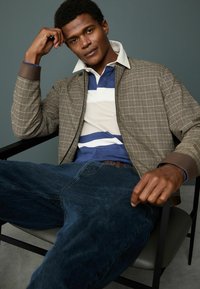 Brown checkered bomber jacket over a blue and white striped polo shirt, paired with dark blue jeans. Smooth textures and simple design.