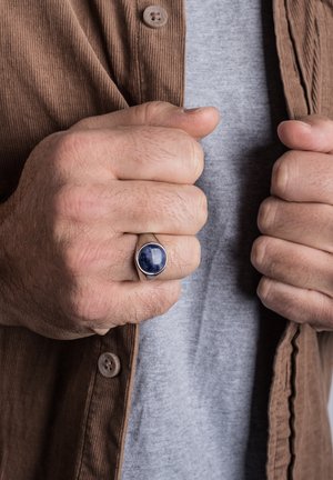 Bague en argent avec une pierre bleu foncé. Poing serré, portant une chemise en velours côtelé marron et un T-shirt gris en dessous. Surface texturée visible.