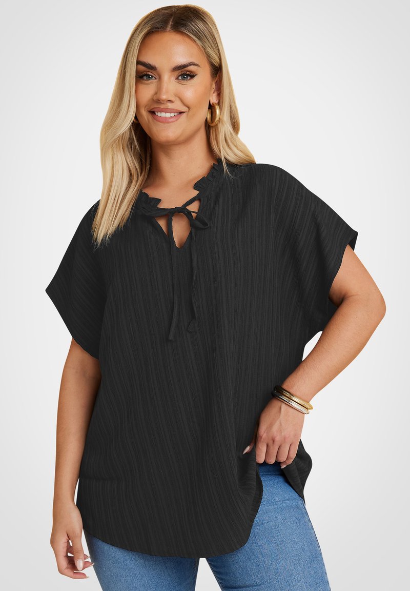 Donna sorridente con capelli biondi lunghi che indossa una blusa nera a maniche corte con scollo a fiocco e jeans blu, accessoriata con braccialetti dorati e orecchini a cerchio.