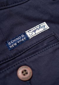 Mörkblå chinos tyg har en vit etikett med blå sömnad som säger "Superdry" och en brun knapp med varumärkets namn.