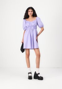 Robe lilas à manches courtes bouffantes avec un corsage smocké et une jupe évasée, accompagnée de chaussures à plateforme noires et d'un sac à main noir.