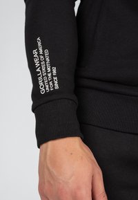 Crna dukserica s rebrastim manžetama. Sadrži bijeli natpis "GORILLA WEAR UNITED STATES OF AMERICA FOR THE MOTIVATED SINCE 1982" na rukavu.
