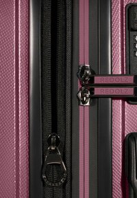 Nahaufnahme eines lila Hartschalenkoffers mit Reißverschluss und doppeltem Schloss, beschriftet mit "REDOLZ", auf einer strukturierten Oberfläche mit schwarzem Rand.