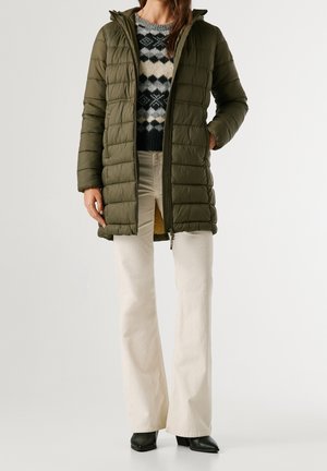 Mujer con chaqueta acolchada con capucha color verde oliva, un suéter negro y blanco con estampado, pantalones crema y botas negras puntiagudas.