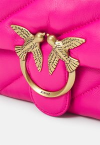 Bolso de piel de color fucsia brillante con un cierre en tono dorado que presenta dos esculturas de pájaros. El bolso tiene una textura acolchada y detalles de costura visibles.