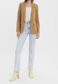 Blazer beige avec un col à revers cranté, associé à un haut côtelé clair, un jean taille haute bleu clair et des bottines à talons carrés jaunes.