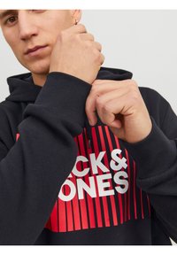 Felpa nera con grande logo rosso e bianco "Jack & Jones", con polsini e cappuccio a coste, indossata da una persona che sta sistemando la manica.
