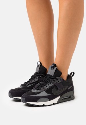 Zapatillas negras con una mezcla de materiales de ante y malla. Cuentan con un destacado acento gris, un cuello alto y una suela de goma texturizada.
