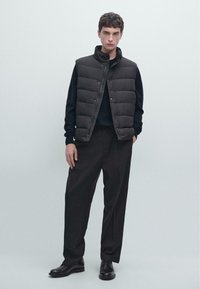 Gilet imbottito nero con cuciture orizzontali, indossato sopra un maglione nero. Pantaloni neri larghi e scarpe nere completano il completo.