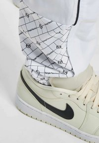 Weißer Jogginghose mit einem schwarzen Karomuster und der Marke "Sergio Tacchini". Graue Sneakers mit einem schwarzen Nike-Swoosh sind teilweise sichtbar.