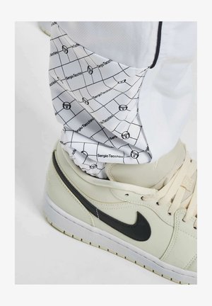 Biely teplákový nohavice s čiernym mriežkovaným vzorom a brandingom "Sergio Tacchini". Šedé tenisky s čiernym logom Nike sú čiastočne viditeľné.