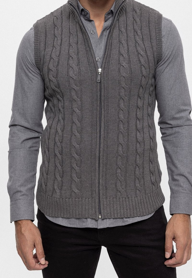 Grijze gebreide vest met een kabelpatroon, voorzien van een rits aan de voorkant en een geribde onderkant, gedragen over een grijs overhemd met knopen.