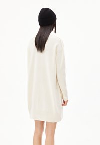 Ecrufarbenes, oversized Strickkleid aus weichem Material mit langen Ärmeln und geripptem Saum. Eine schwarze Strickmütze bildet einen Kontrast zum Outfit.